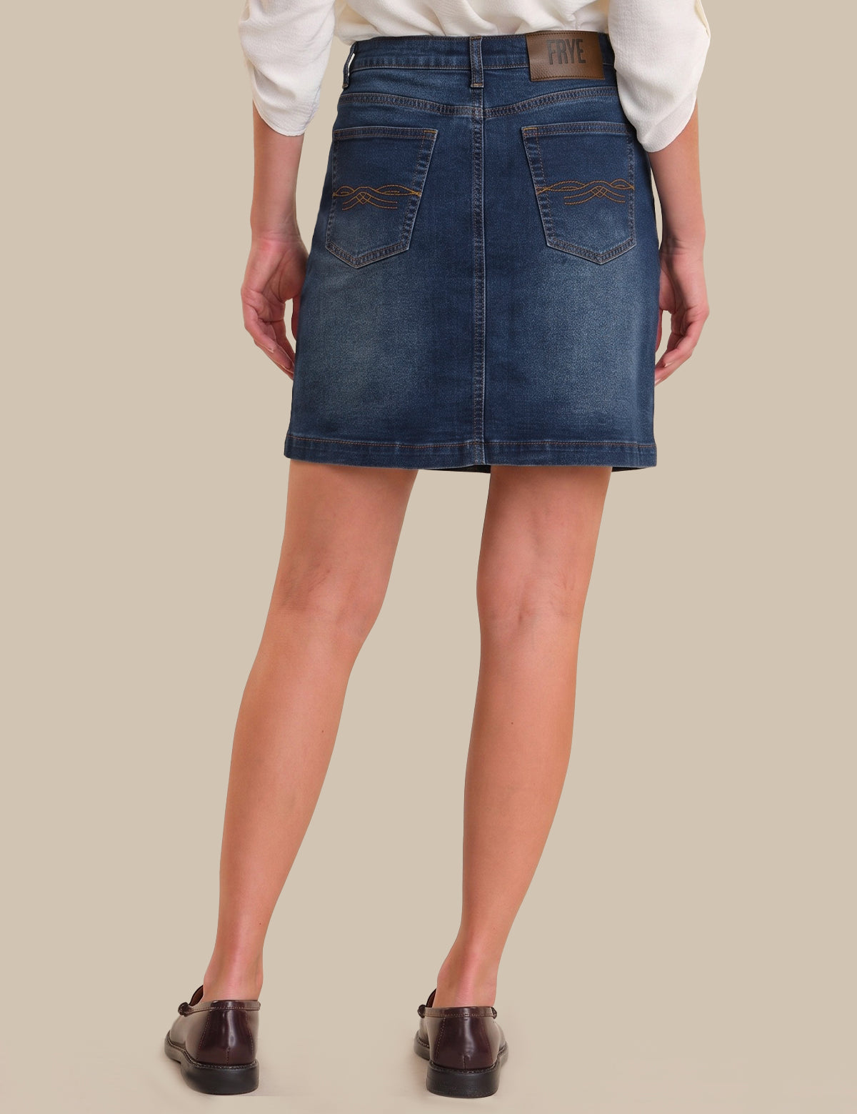 A model from the back wearing a blue denim mini skirt with contrast stitching and back pockets, paired with a white long-sleeve top and dark brown leather loafers, against a solid beige background.