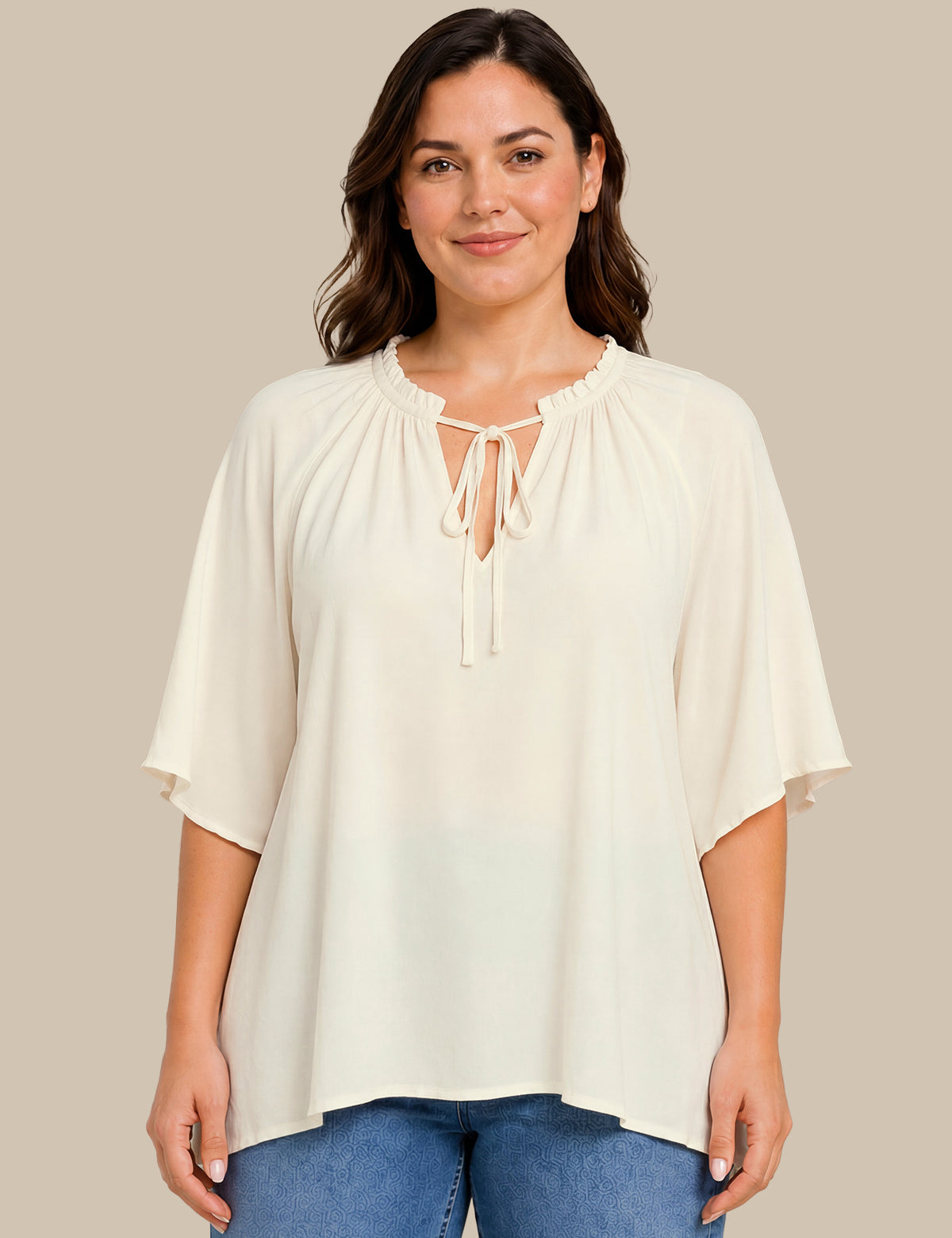 A woman wearing a cream-colored short-sleeve blouse with a tie-neck detail, paired with blue jeans, standing against a solid beige background.