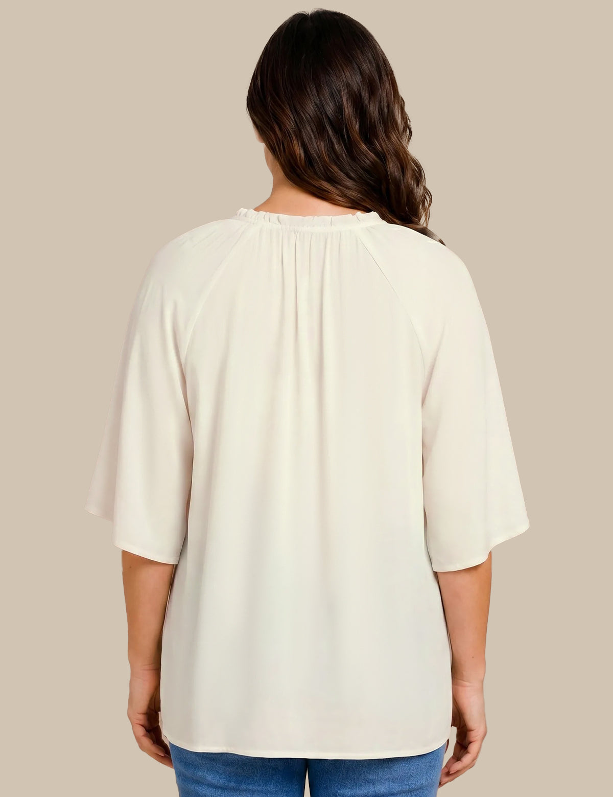 A woman viewed from the back wearing a cream-colored short-sleeve top with a gathered neckline and blue jeans, set against a solid beige background.