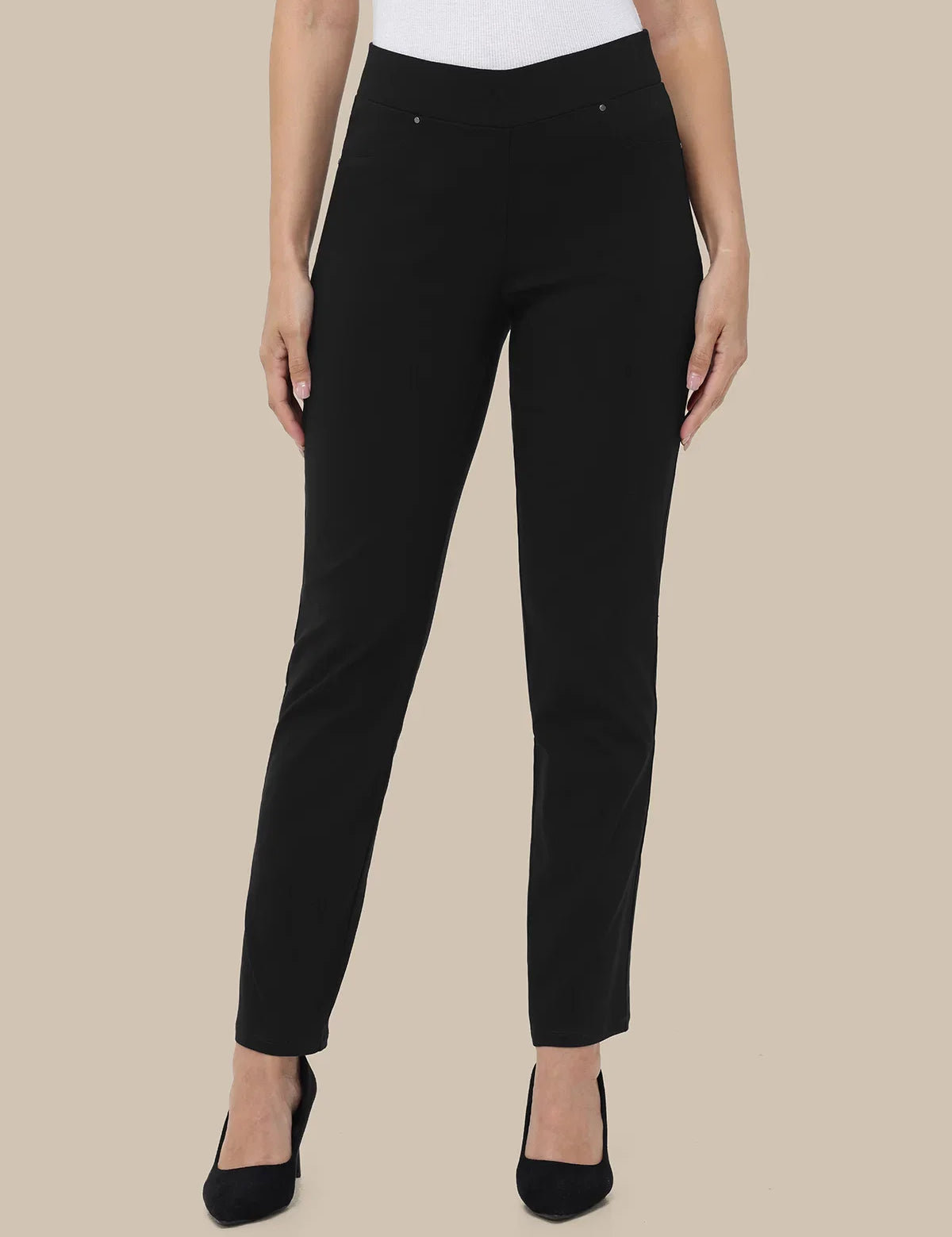 A model wearing slim-fit black trousers with a white top and black pointed-toe heels, shown against a solid beige background.