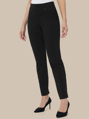 A model wearing slim-fit black ankle-length pants with a white top and black pointed-toe heels, shown against a plain beige studio background.