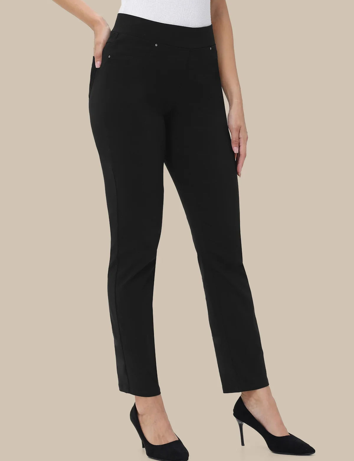 A model wearing black slim-fit ankle-length trousers with side pockets, paired with a white top and black pointed-toe heels, shown against a solid beige background.