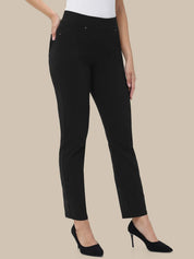A model wearing black slim-fit ankle-length trousers with side pockets, paired with a white top and black pointed-toe heels, shown against a solid beige background.