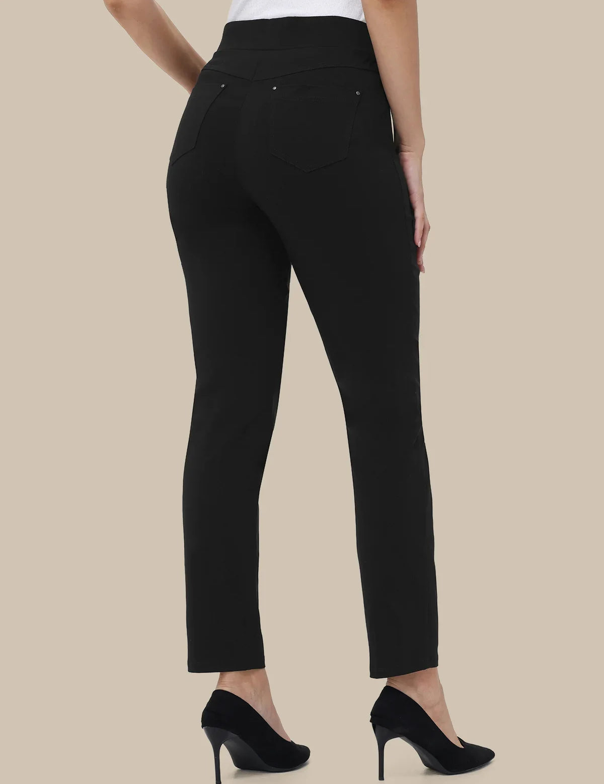 Black slim-fit ankle-length pants shown on a model from the back, paired with a white top and black high heels, against a solid beige background.