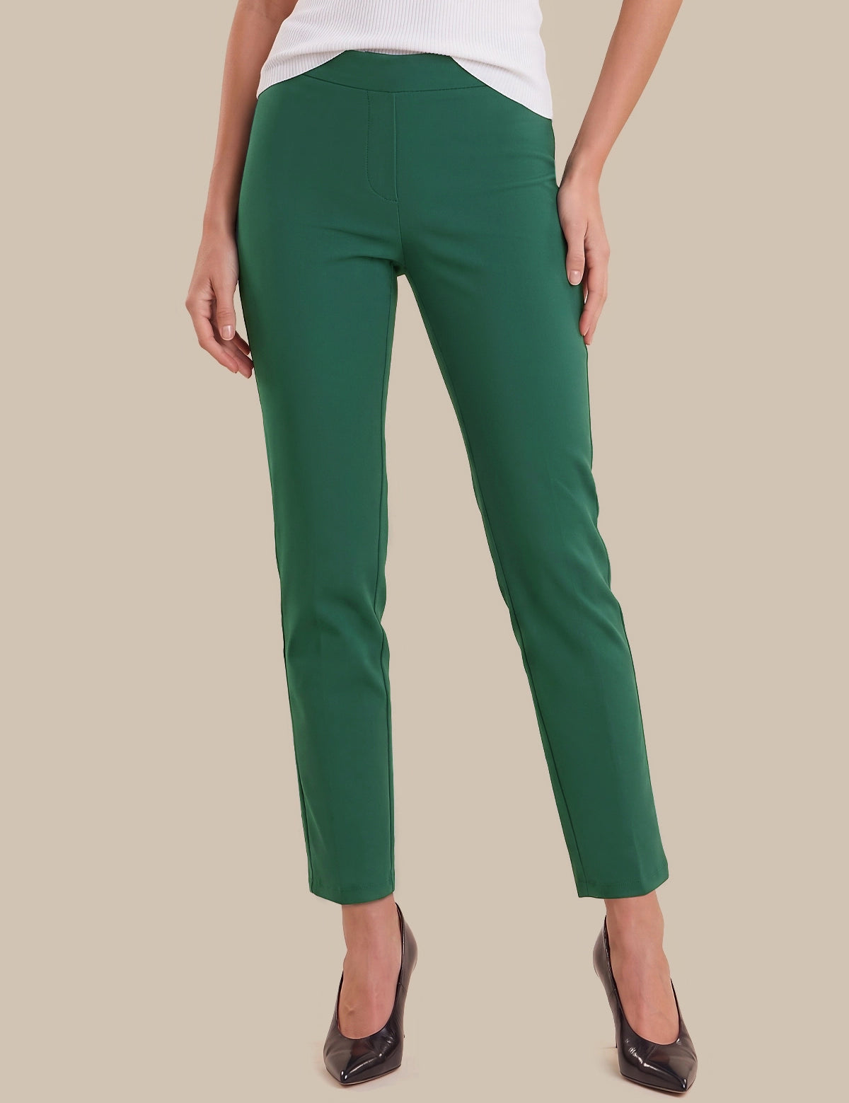 A model wearing a pair of slim-fit, ankle-length green trousers with a white top and black pointed-toe heels, shown from the waist down against a solid beige background.