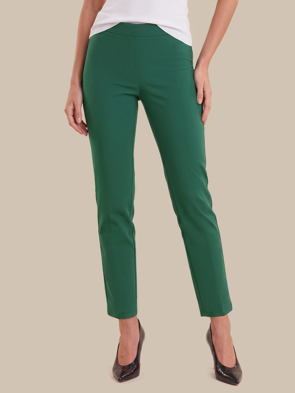 A model wearing a pair of slim-fit, ankle-length green trousers with a white top and black pointed-toe heels, shown from the waist down against a solid beige background.