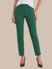 A model wearing a pair of slim-fit, ankle-length green trousers with a white top and black pointed-toe heels, shown from the waist down against a solid beige background.