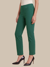 A model wearing a pair of slim-fit, ankle-length green trousers with a white top and black patent leather pumps, shown against a solid beige background.