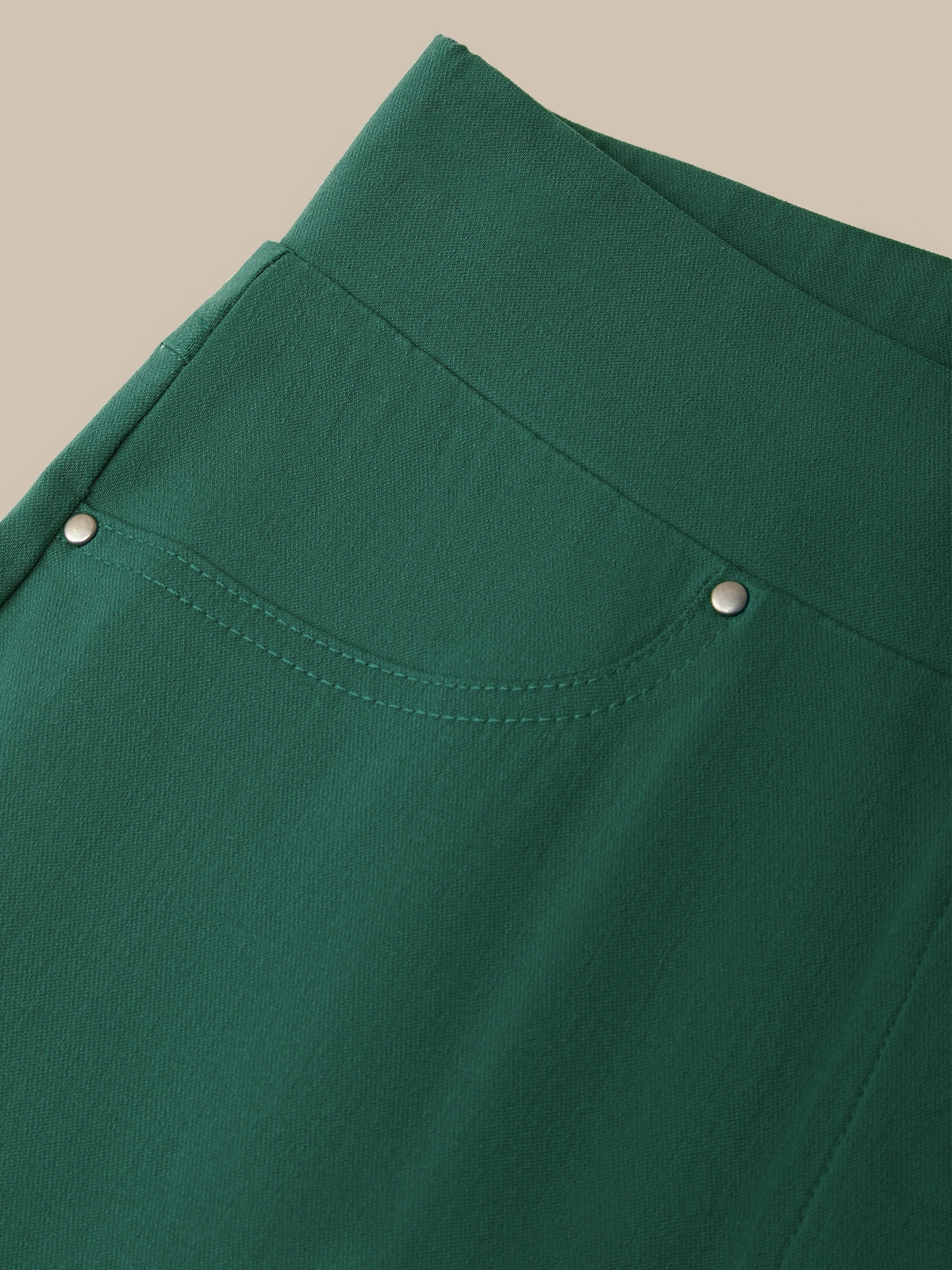 Close-up of a dark green garment, likely a skirt or dress, showing a curved seam, a decorative pocket with silver-tone snap details, and a clean beige background.