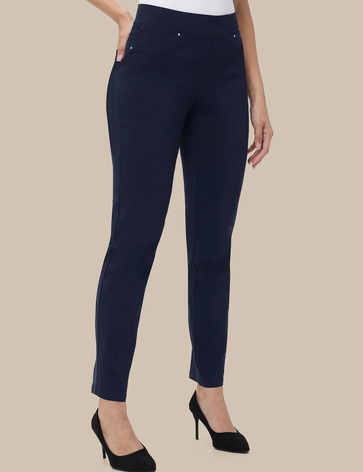 A model wearing navy blue slim-fit ankle-length trousers with a white top and black high heels, shown against a solid beige background.