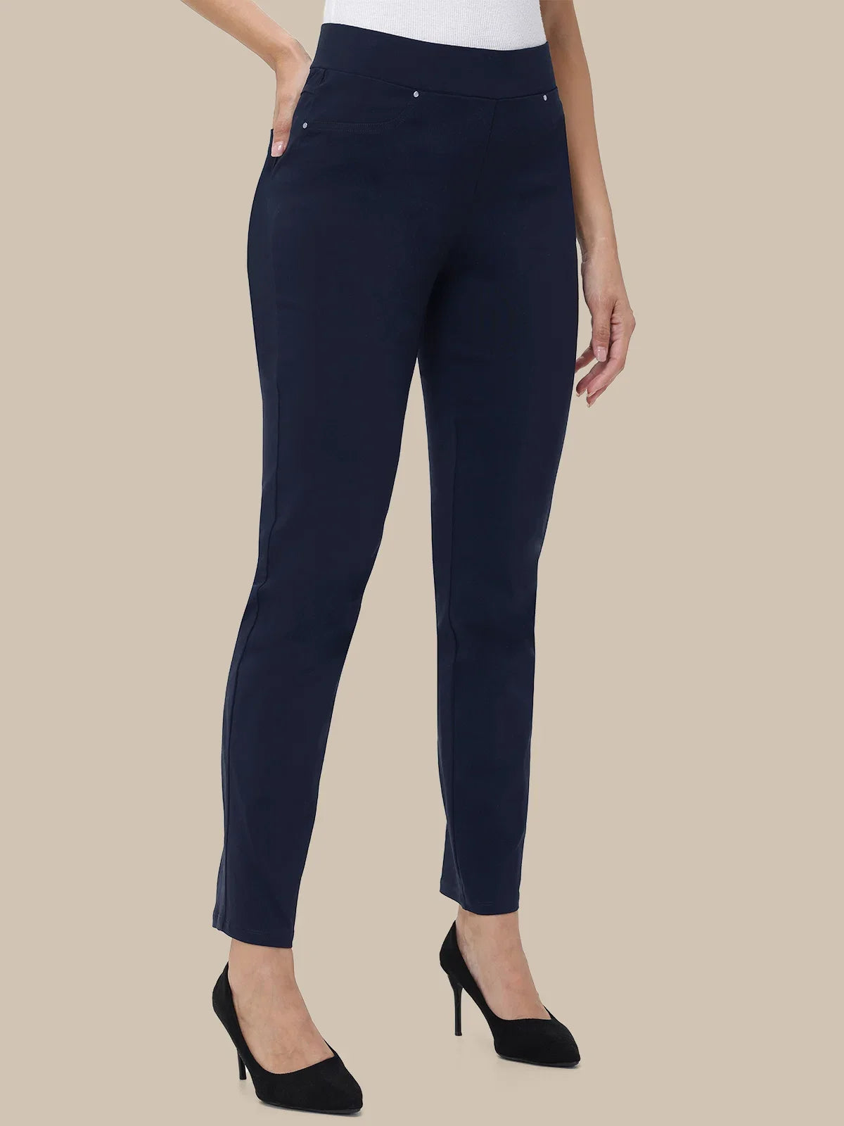 A model wearing navy blue slim-fit ankle-length trousers with a white top and black high heels, shown against a solid beige background.