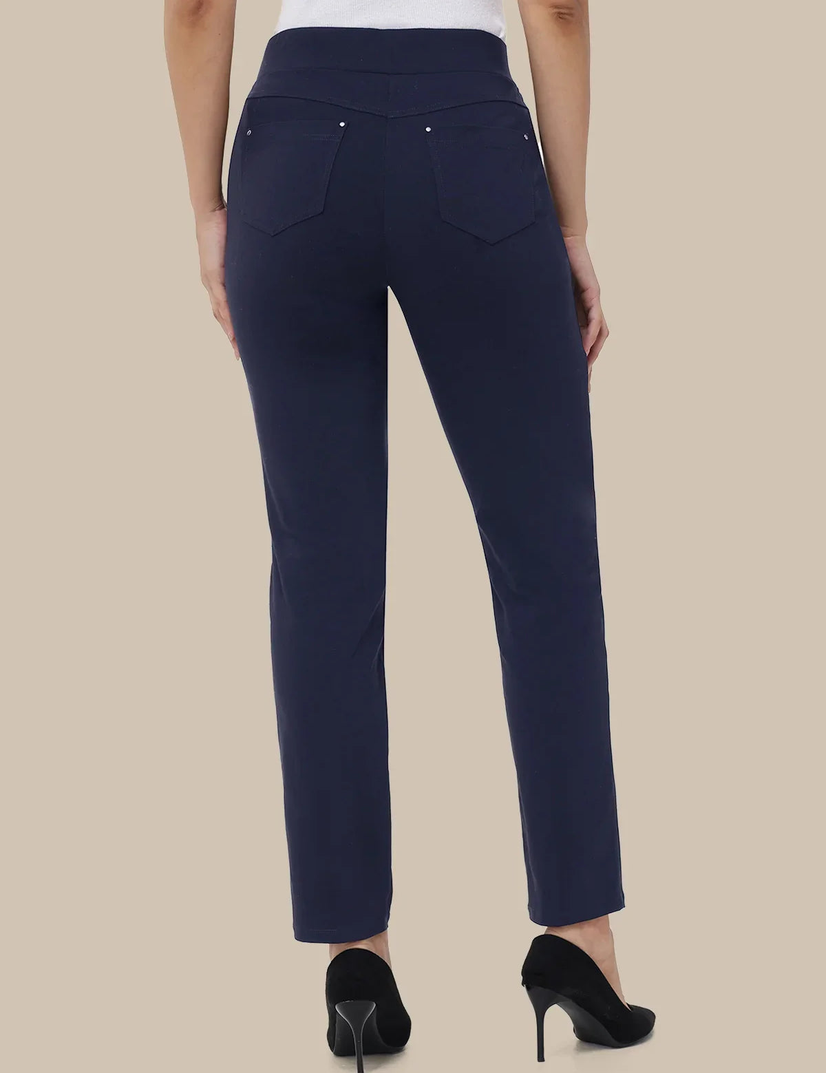 A person shown from the back wearing navy blue high-waisted straight-leg pants with silver button details on the back pockets, paired with black high heels against a solid beige background.