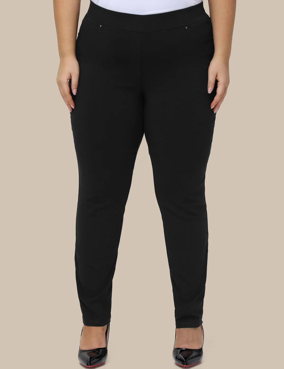 A model wearing black slim-fit pants with a wide elastic waistband, paired with a white top and black pointed-toe heels, shown against a solid beige background.