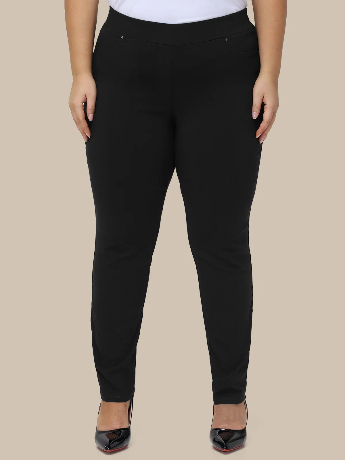 A model wearing black slim-fit pants with a wide elastic waistband, paired with a white top and black pointed-toe heels, shown against a solid beige background.