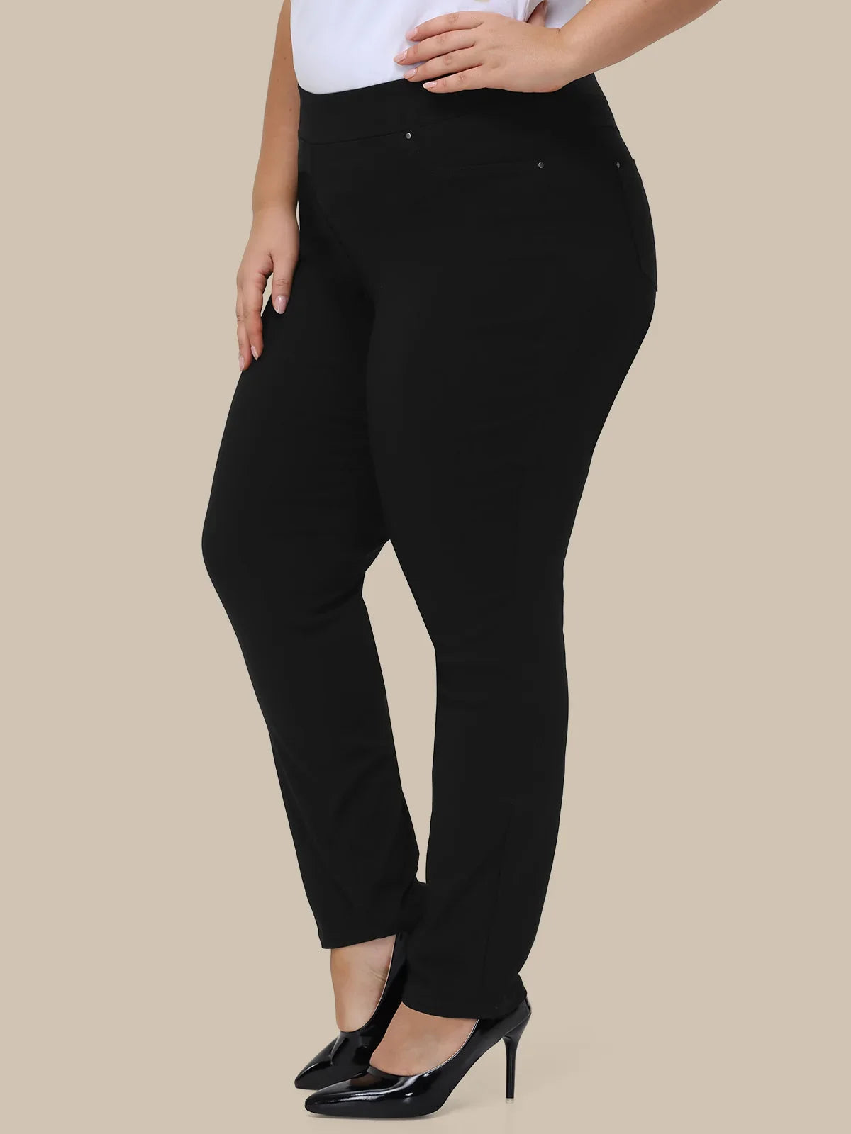 A model wearing black slim-fit pants with a white top and black patent leather heels, shown from the waist down against a solid beige background.