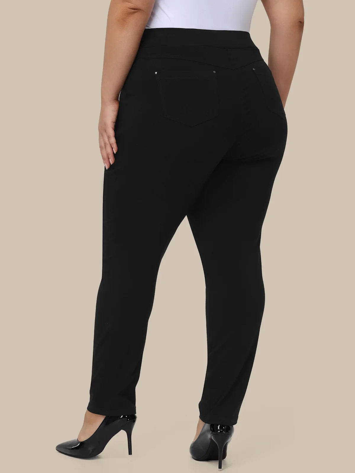 A model shown from the back wearing black high-waisted slim-fit pants with a white top and black heels, set against a solid beige background.