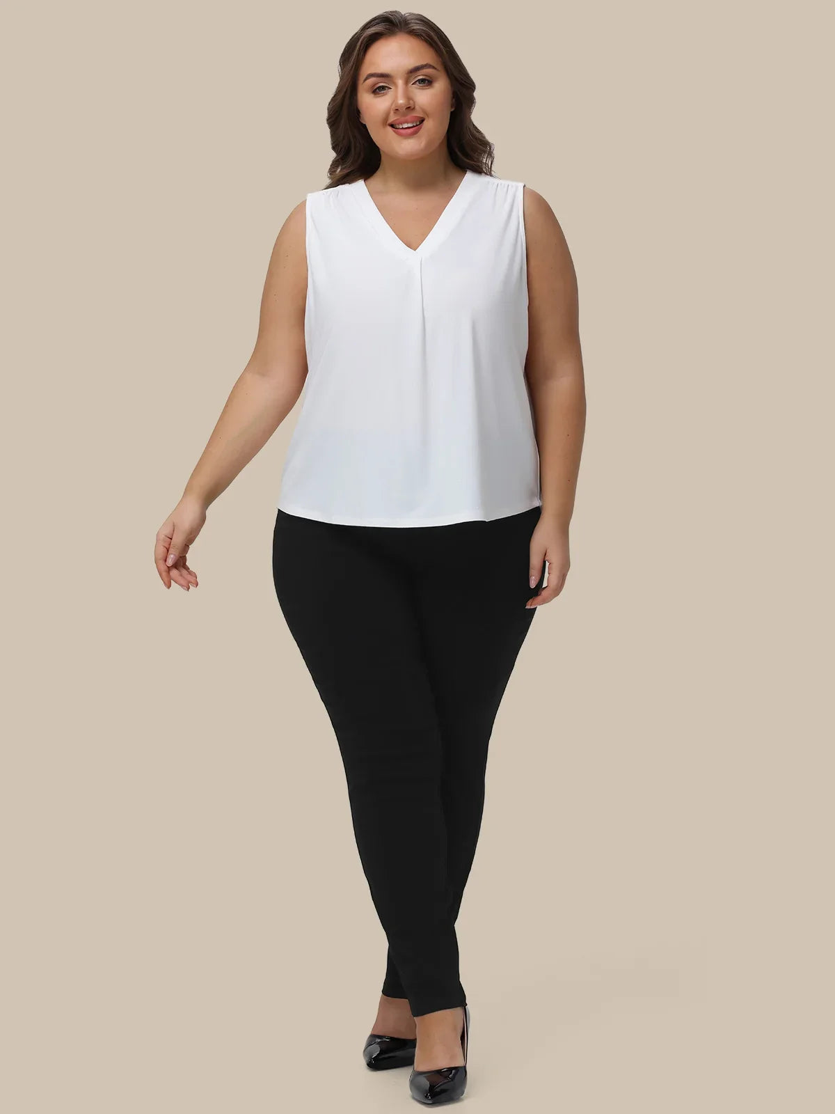 A woman modeling a sleeveless white V-neck top paired with black pants and black heels, standing against a solid beige background.