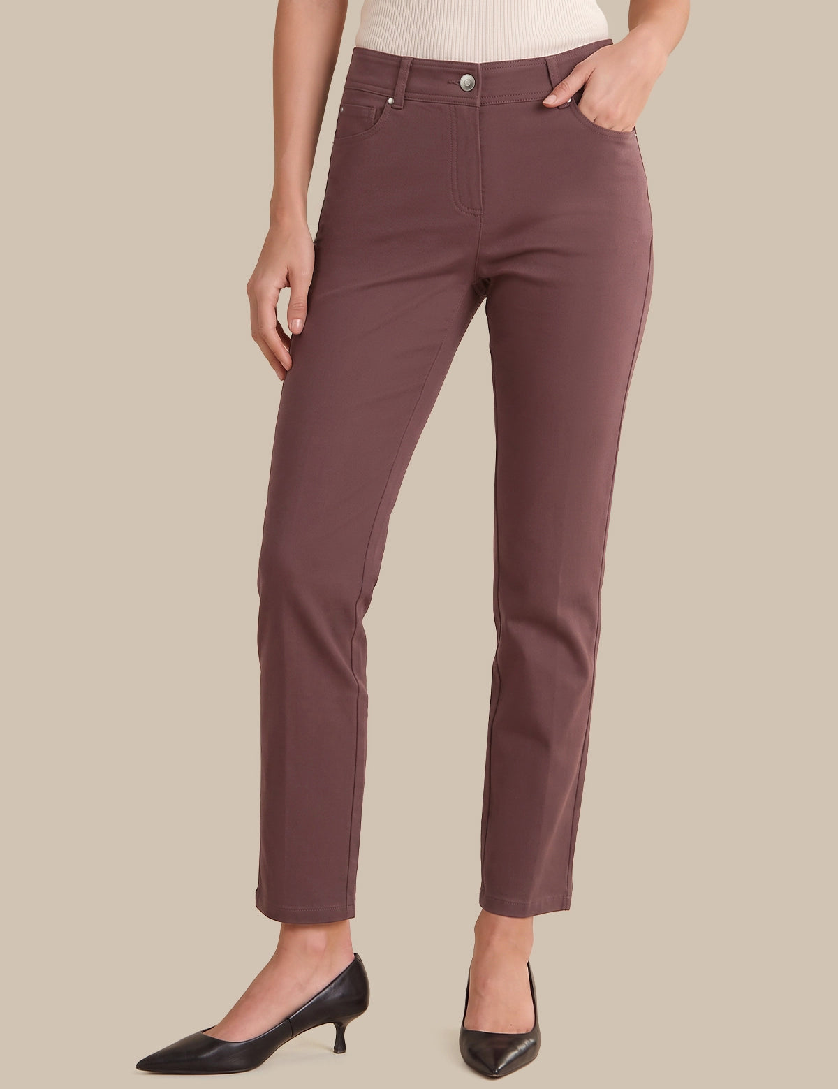 A model wearing slim-fit, ankle-length trousers in a muted dusty rose or mauve color, paired with black pointed-toe heels and a light-colored top, set against a solid beige background.