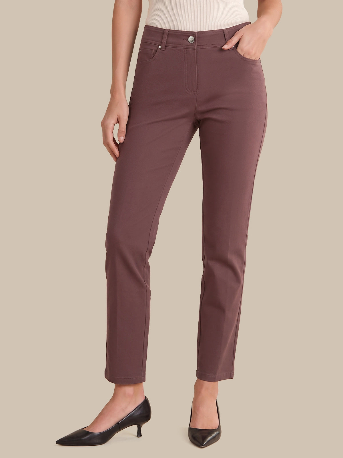 A model wearing slim-fit, ankle-length trousers in a muted dusty rose or mauve color, paired with black pointed-toe heels and a light-colored top, set against a solid beige background.