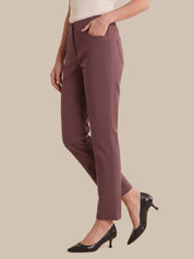 A model wearing a pair of straight-leg, ankle-length trousers in a dusty mauve-brown color, paired with black pointed-toe heels and a cream top, set against a solid beige background.