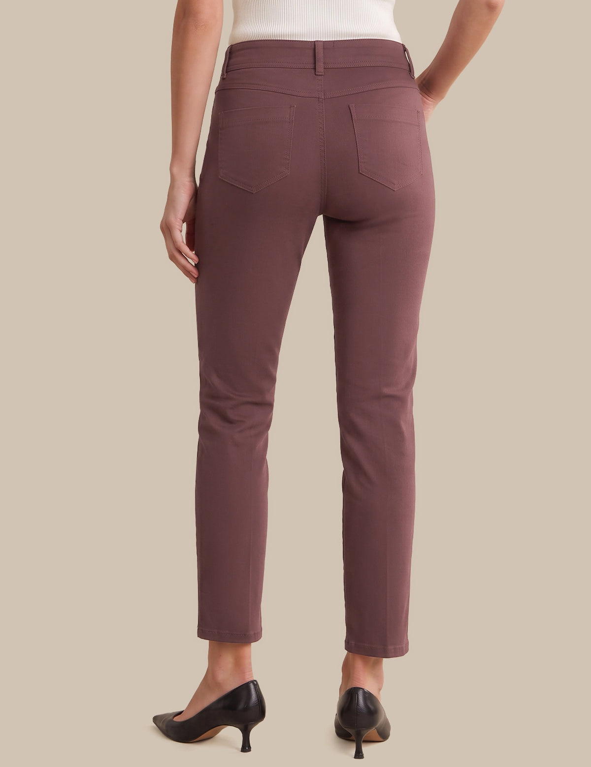 Rear view of a model wearing slim-fit ankle-length pants in a muted dusty rose or mauve color, paired with black pointed-toe heels and a white top, against a solid beige background.