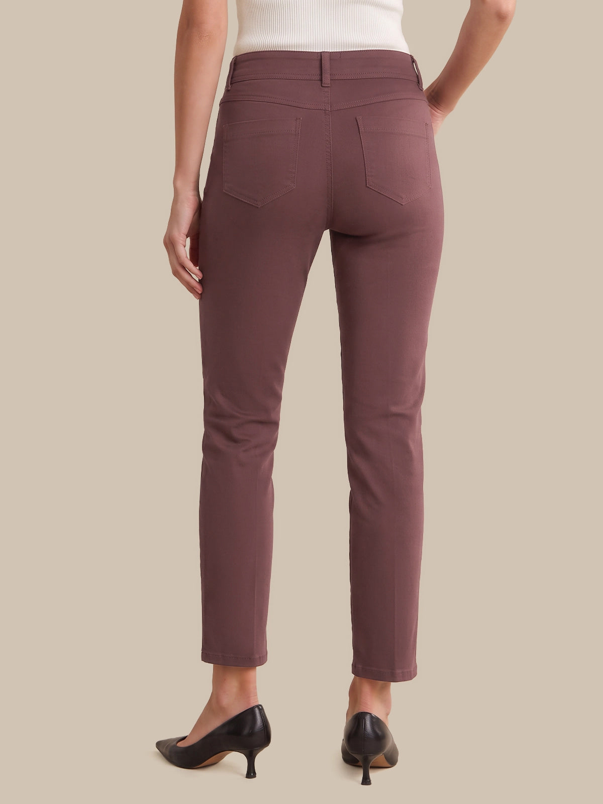 Rear view of a model wearing slim-fit ankle-length pants in a muted dusty rose or mauve color, paired with black pointed-toe heels and a white top, against a solid beige background.