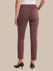 Rear view of a model wearing slim-fit ankle-length pants in a muted dusty rose or mauve color, paired with black pointed-toe heels and a white top, against a solid beige background.