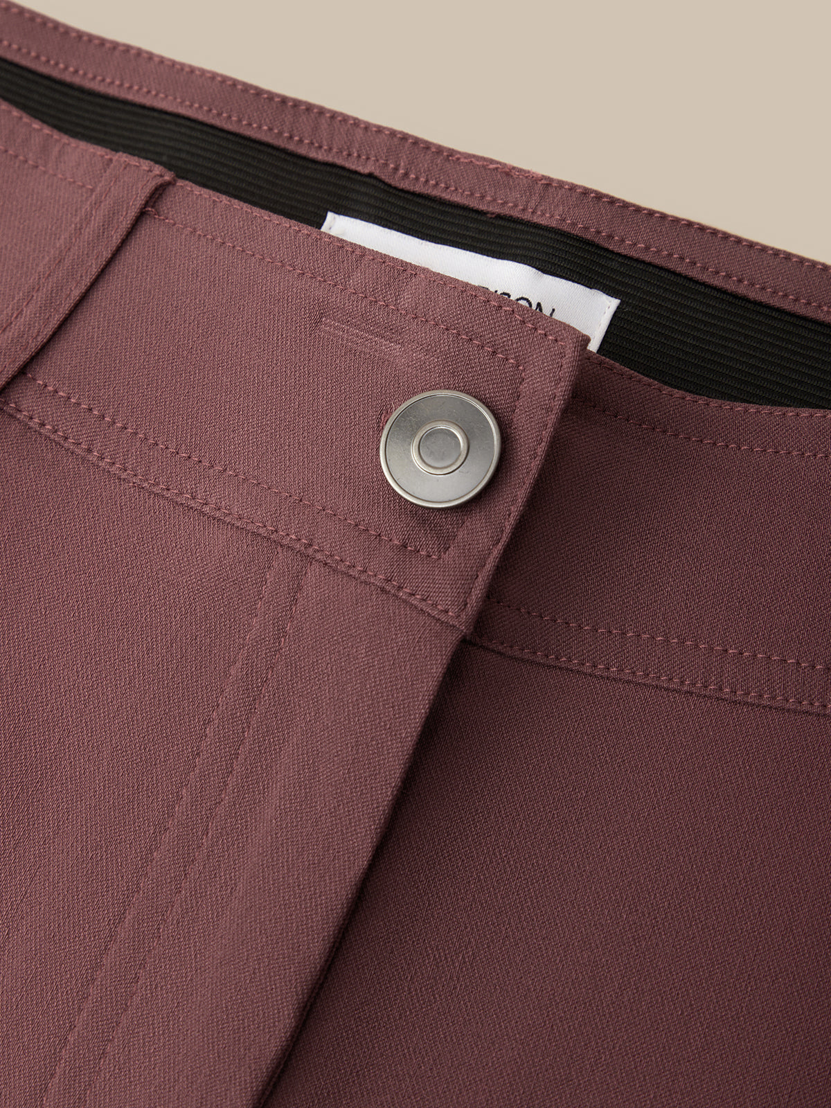 Close-up detail of mauve-brown trousers showing a button closure, waistband, and fabric texture against a neutral beige background.
