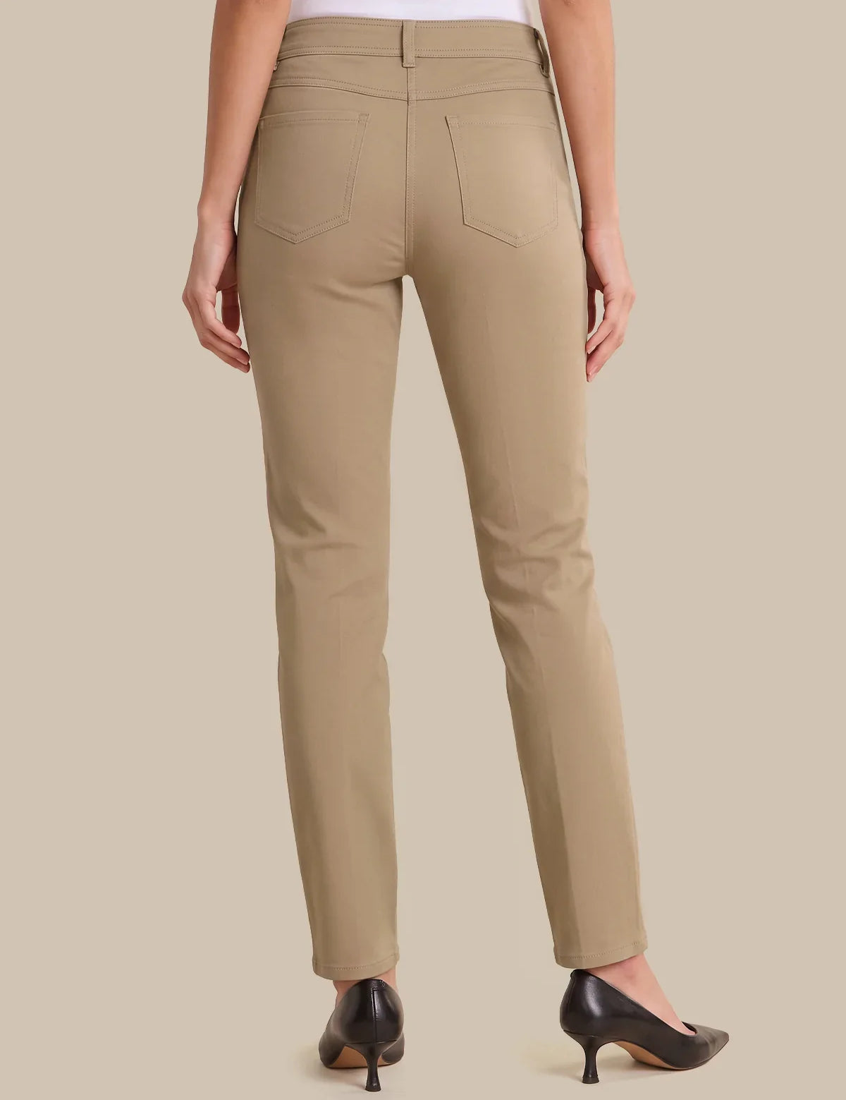 A person shown from the back wearing straight-leg beige pants, a white top, and black pointed-toe heels against a plain beige studio background.