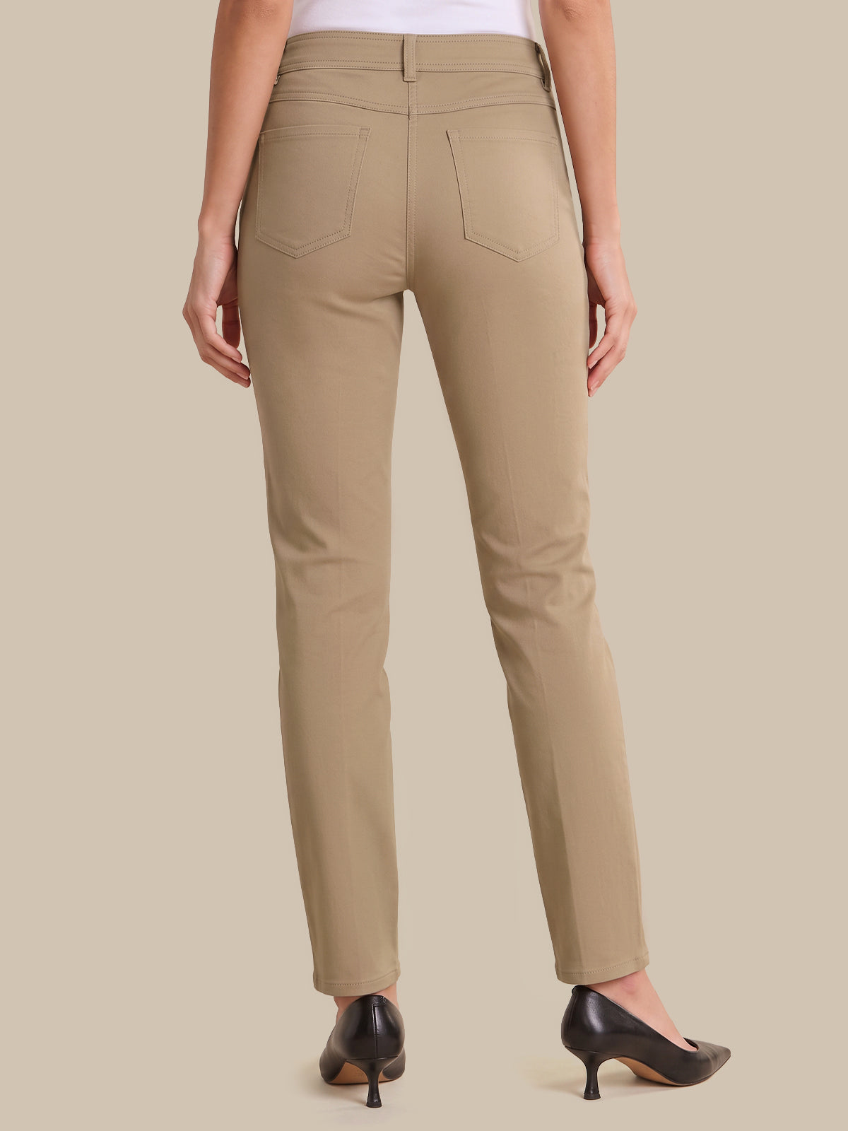 A person shown from the back wearing straight-leg beige pants, a white top, and black pointed-toe heels against a plain beige studio background.