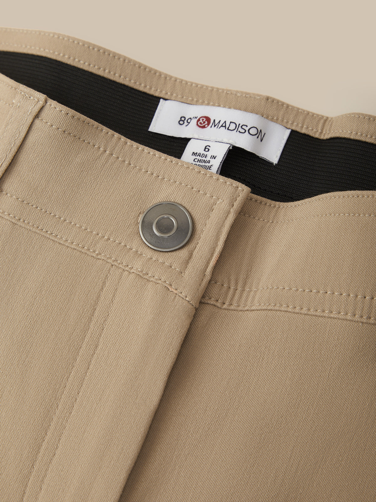 Close-up of beige trousers showing the waistband, a silver metal button, and a white brand label reading 89 Madison.