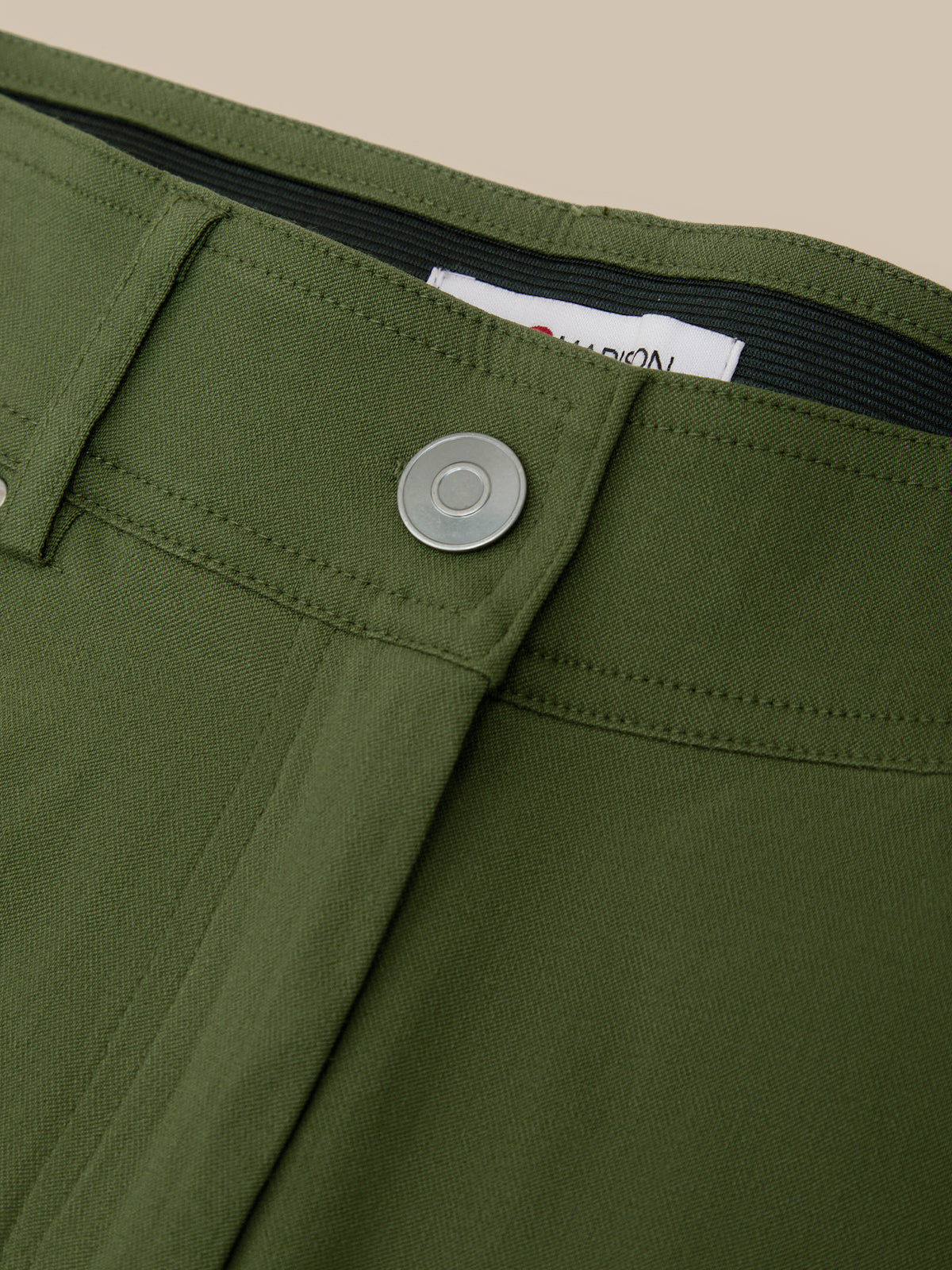 Close-up of olive green trousers showing the waistband, button closure, belt loops, and inner label on a plain beige background.