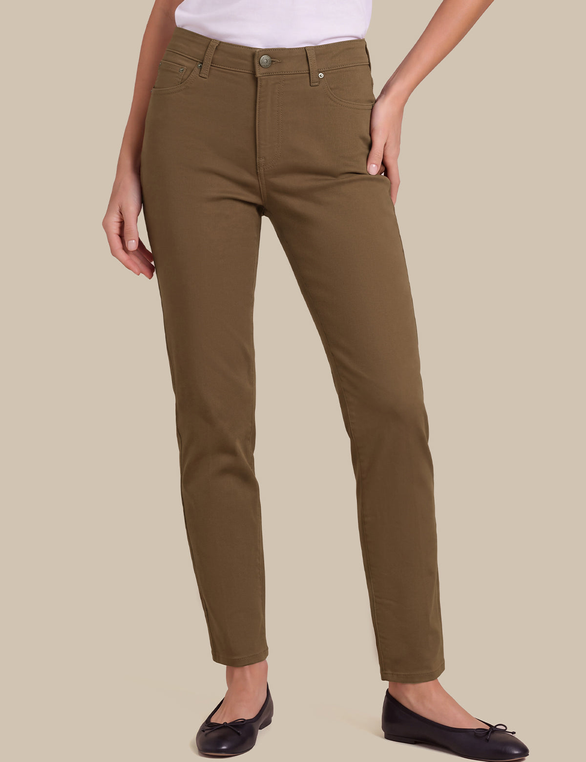 A person wearing slim-fit olive brown pants with a white top and black flats, shown from the waist down against a plain beige studio background.