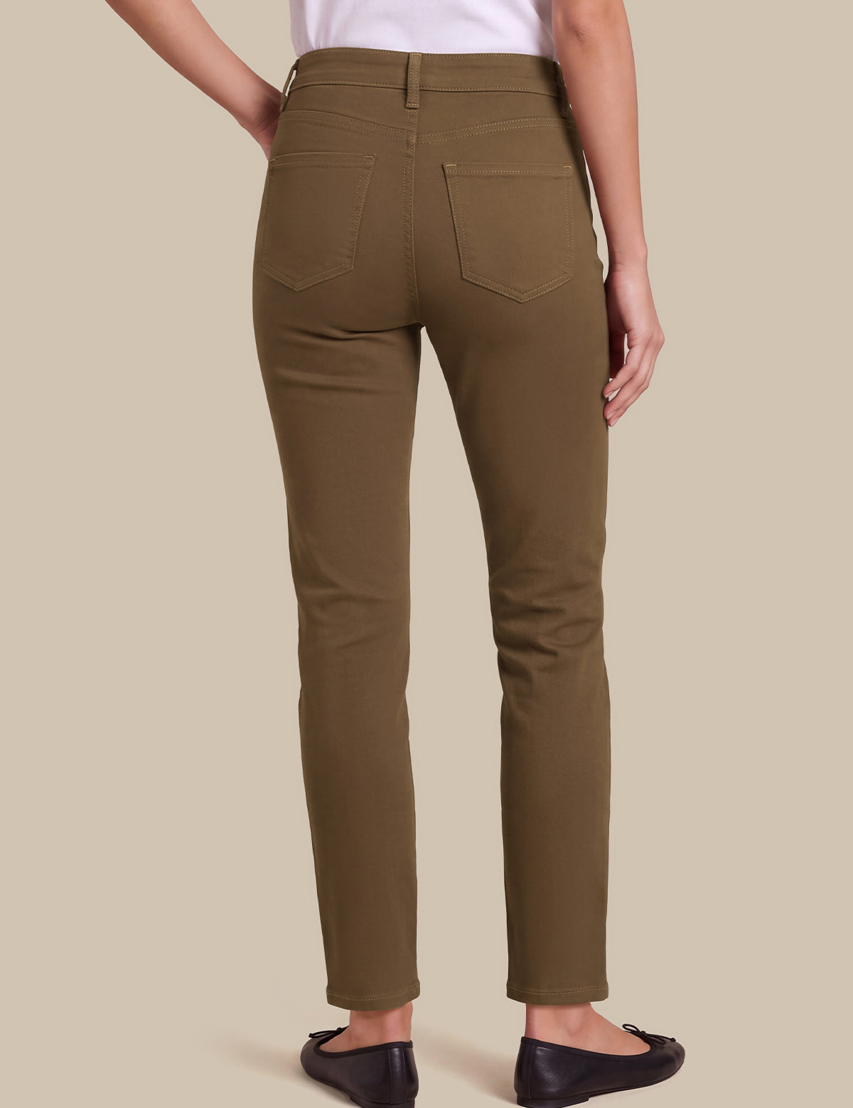 A person shown from the back wearing slim-fit olive green pants, a white top, and black flats against a plain beige studio background.