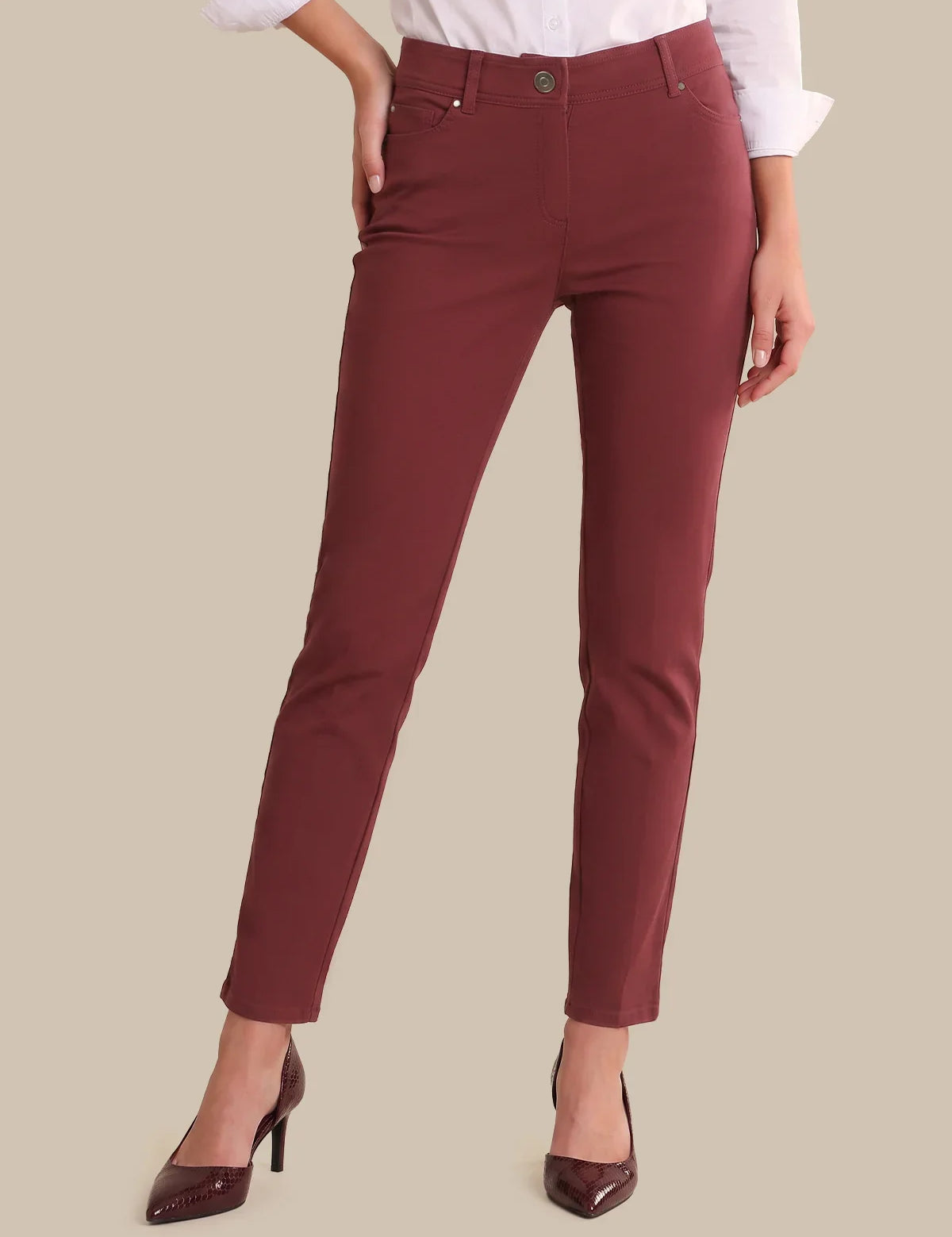 A model wearing a pair of slim-fit, high-waisted ankle-length pants in a deep burgundy red. The image is cropped from the waist down, showing the model wearing a white collared shirt and burgundy pointed-toe heels against a solid beige background.