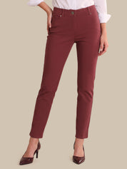 A model wearing a pair of slim-fit, high-waisted ankle-length pants in a deep burgundy red. The image is cropped from the waist down, showing the model wearing a white collared shirt and burgundy pointed-toe heels against a solid beige background.