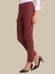 A model wearing slim-fit, ankle-length trousers in a deep burgundy color, styled with a white long-sleeve blouse and dark brown patent leather pumps, set against a solid beige background.