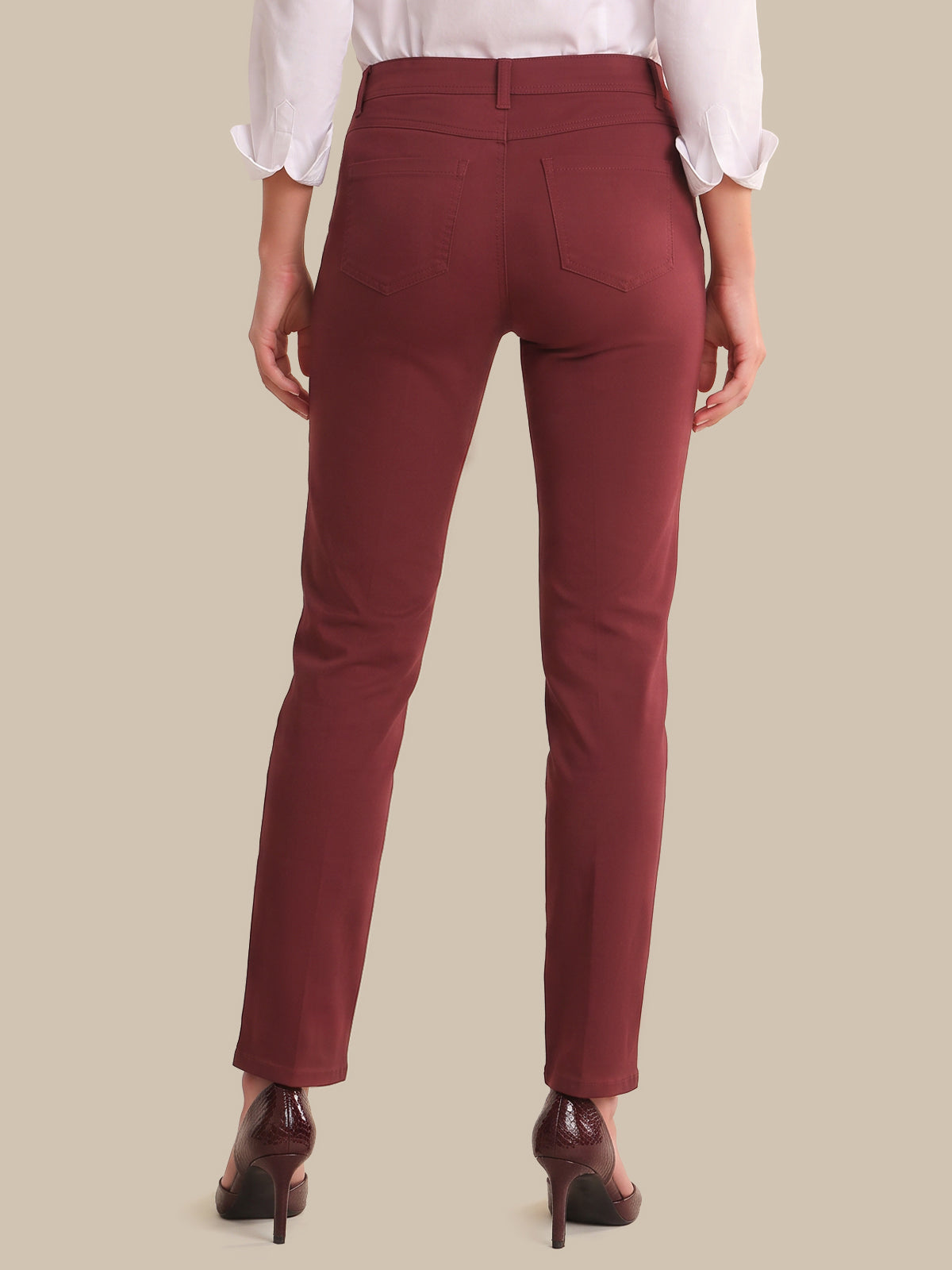 A person shown from the back wearing slim-fit burgundy trousers, a white blouse with rolled sleeves, and dark brown high heels against a plain beige studio background.