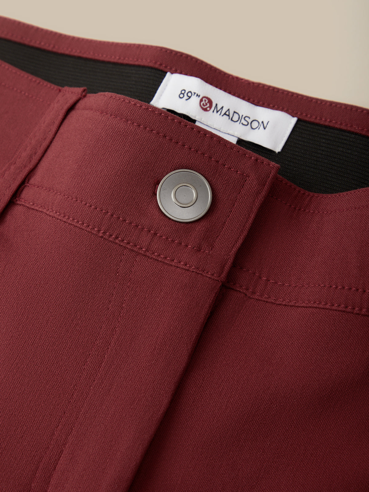 Close-up of burgundy trousers showing the waistband, a silver metal button, and a white interior label reading 89th & MADISON.