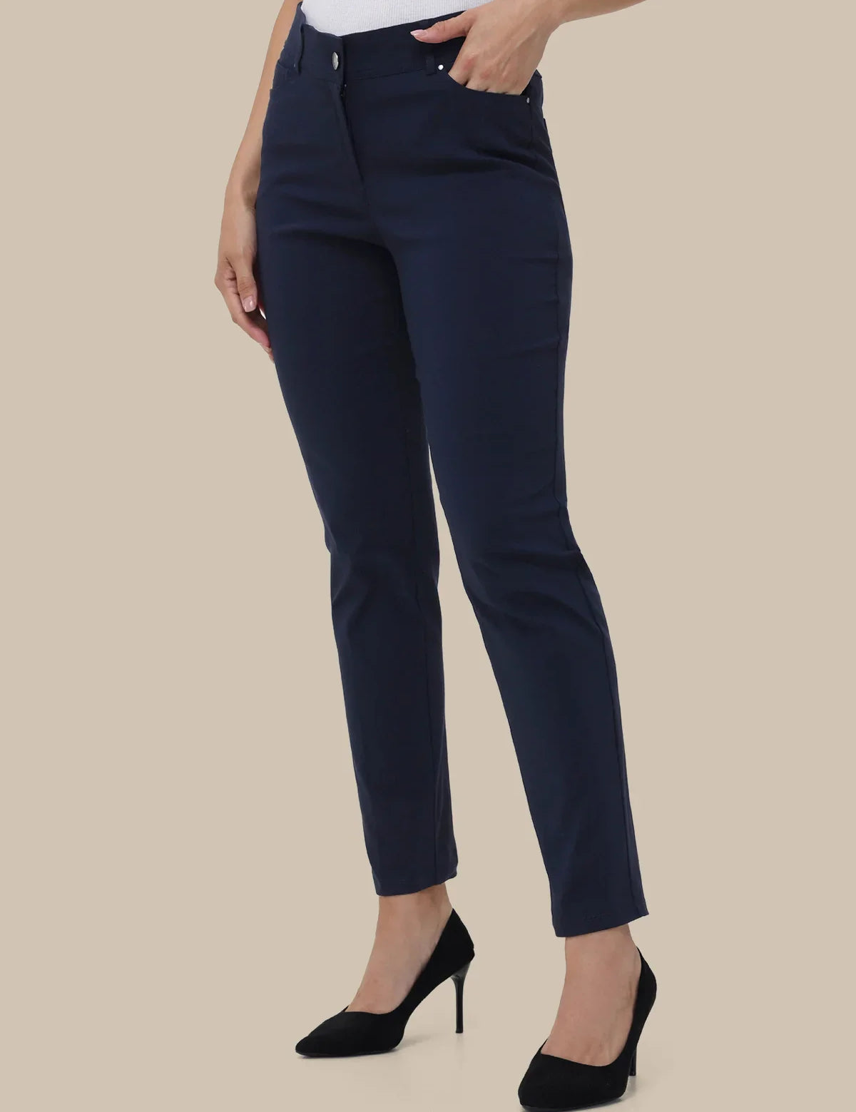 A model wearing navy blue slim-fit ankle-length trousers, styled with a white top and black high heels against a solid beige background.