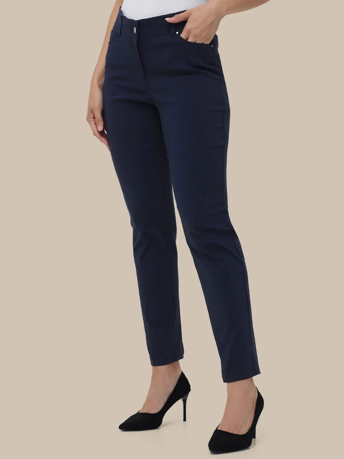 A model wearing navy blue slim-fit ankle-length trousers, styled with a white top and black high heels against a solid beige background.