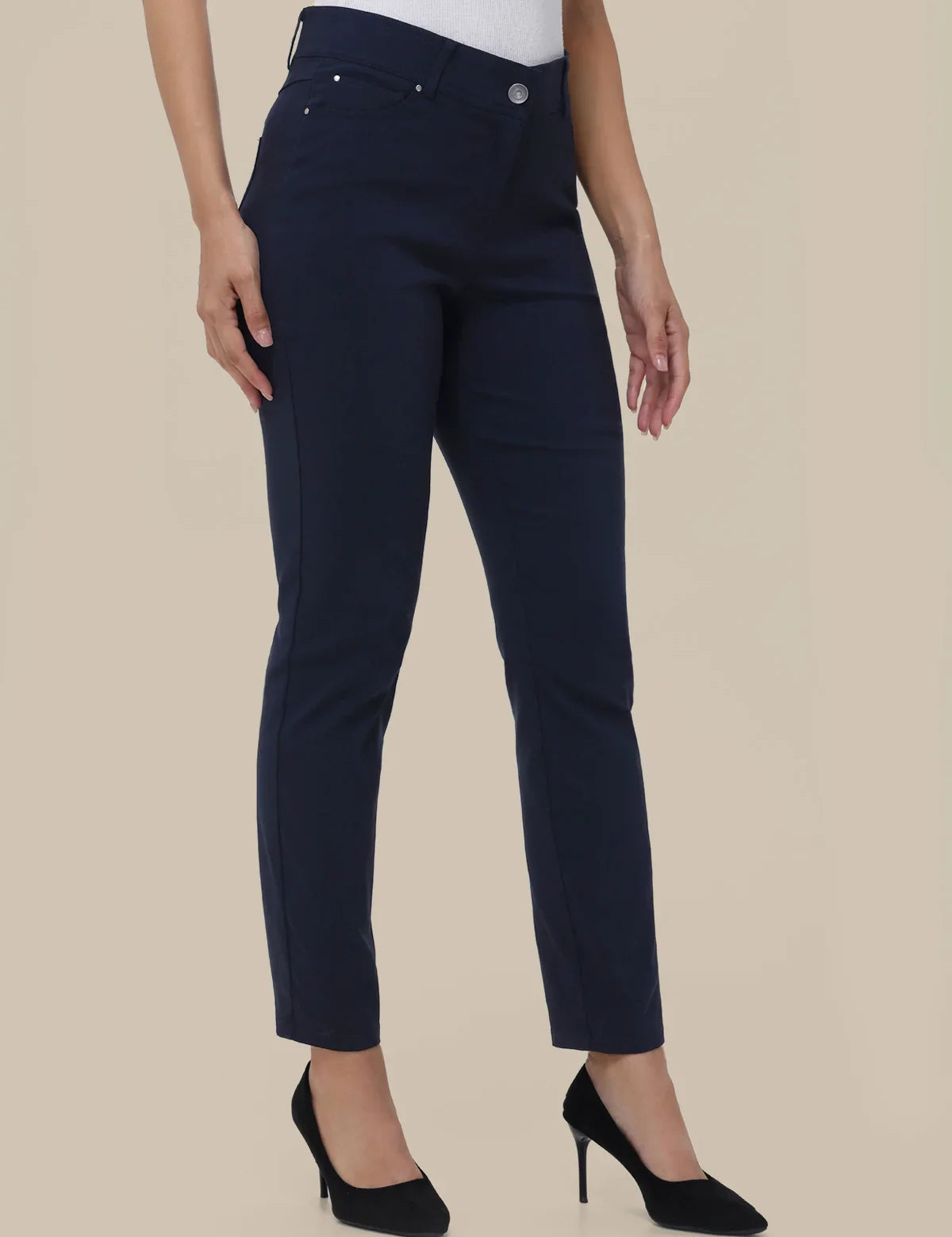 A model wearing a pair of dark navy blue slim-fit ankle-length jeans with a white top and black high heels, shown from the waist down against a solid beige background.