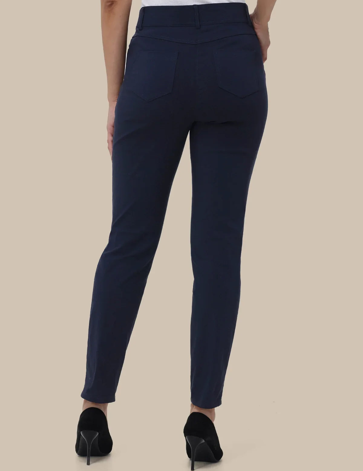 A person shown from the back wearing slim-fit navy blue pants, a white top, and black high heels against a plain beige studio background.