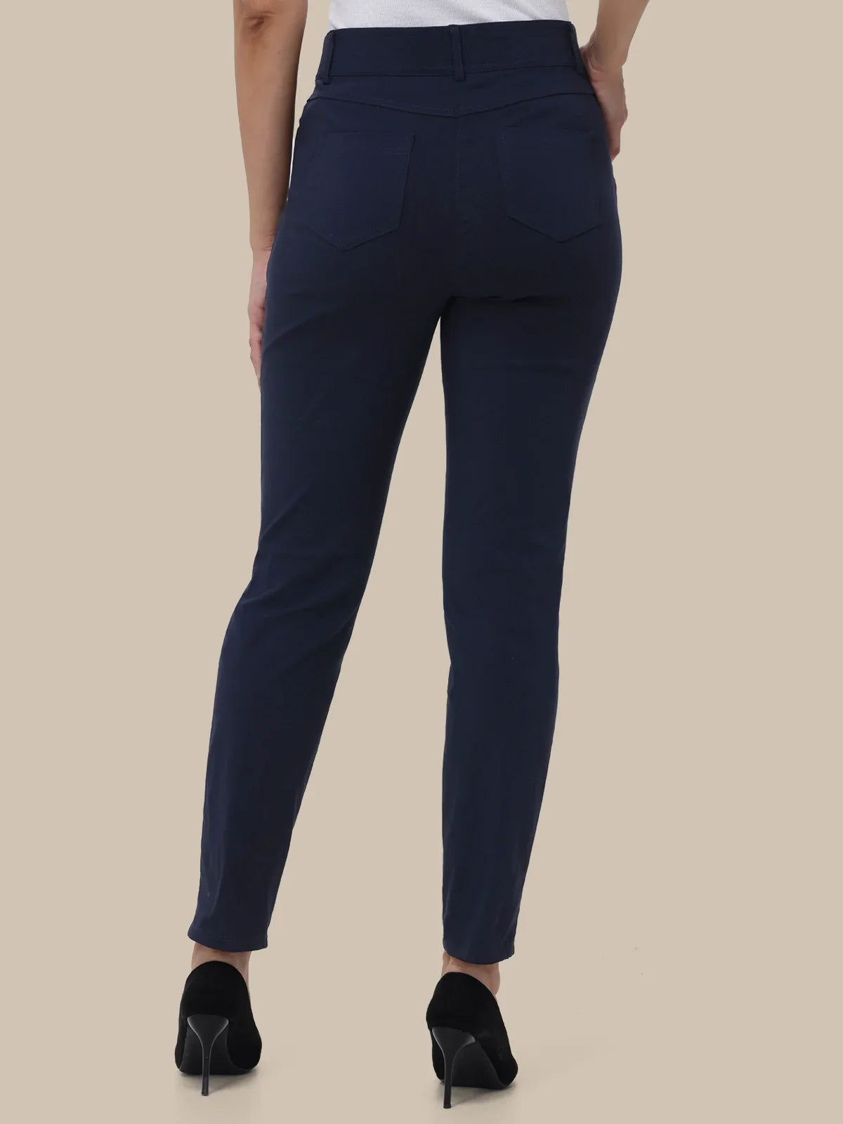 A person shown from the back wearing slim-fit navy blue pants, a white top, and black high heels against a plain beige studio background.