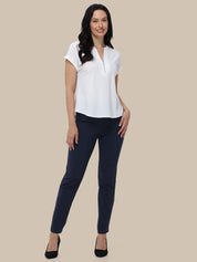 A woman modeling a white short-sleeve top with a split V-neckline and dark navy pants, standing against a solid beige background.
