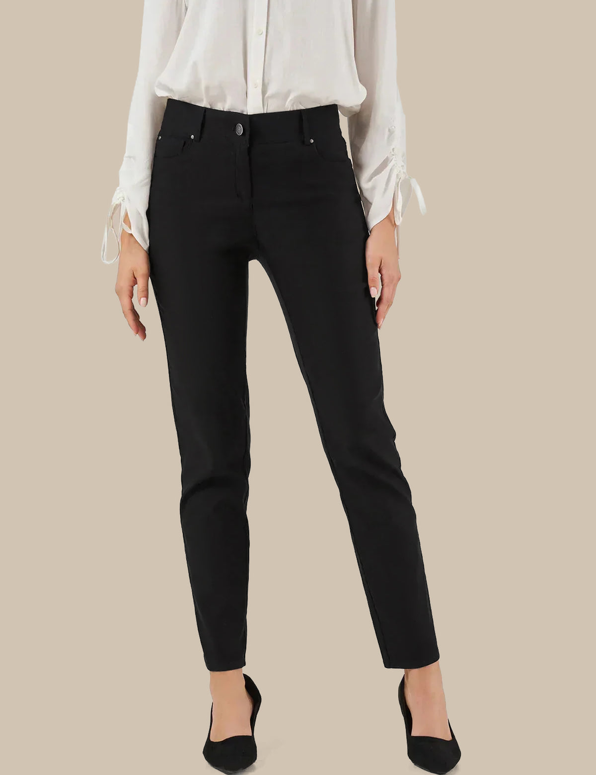 A model wearing slim-fit black ankle-length jeans with a white long-sleeve blouse and black pointed-toe heels, shown against a solid beige background.
