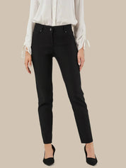 A model wearing slim-fit black ankle-length jeans with a white long-sleeve blouse and black pointed-toe heels, shown against a solid beige background.