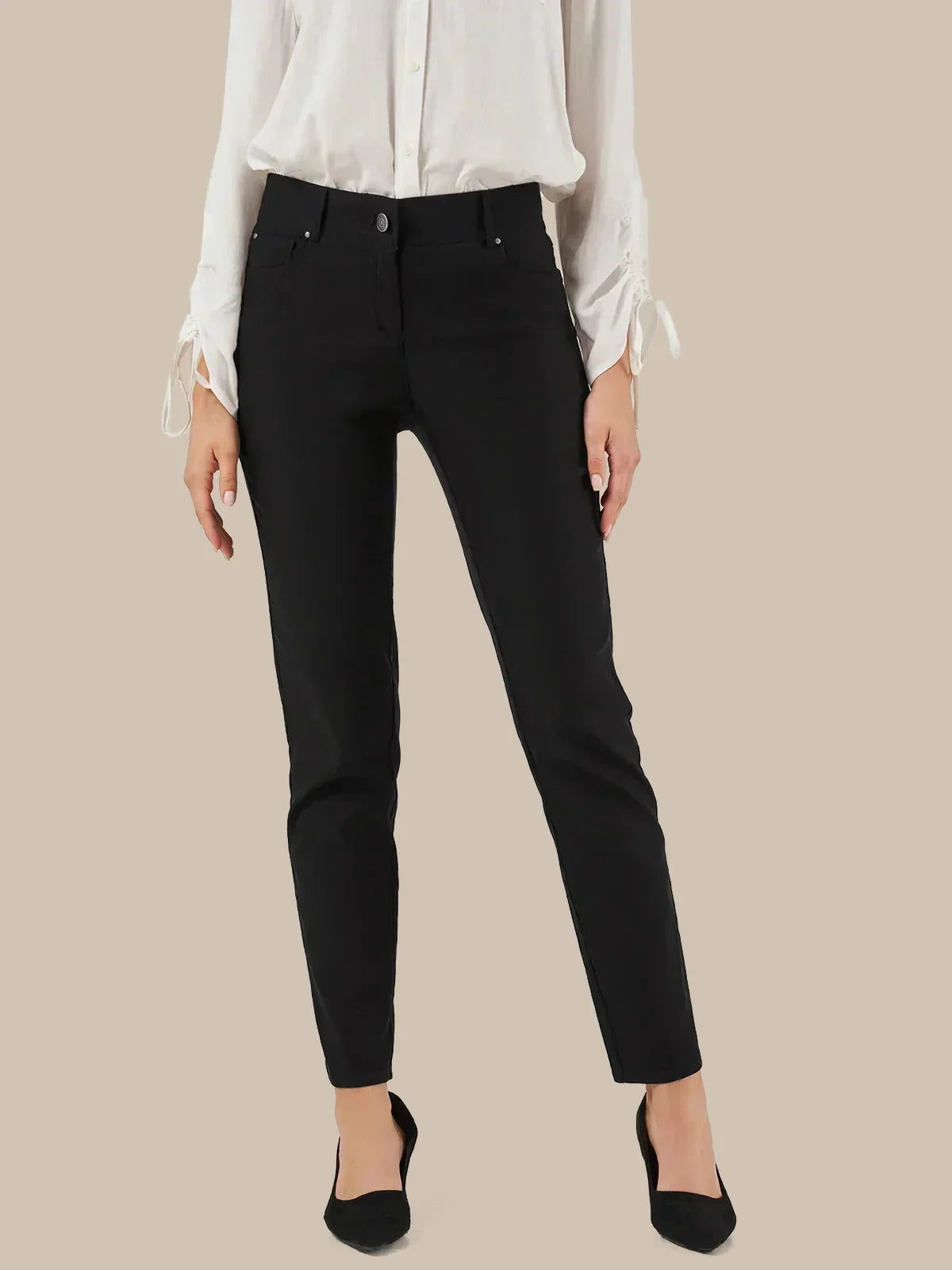 A model wearing slim-fit black ankle-length jeans with a white long-sleeve blouse and black pointed-toe heels, shown against a solid beige background.