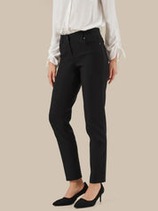 A model wearing black slim-fit ankle-length trousers, styled with a white long-sleeve blouse and black pointed-toe heels, against a solid beige background.