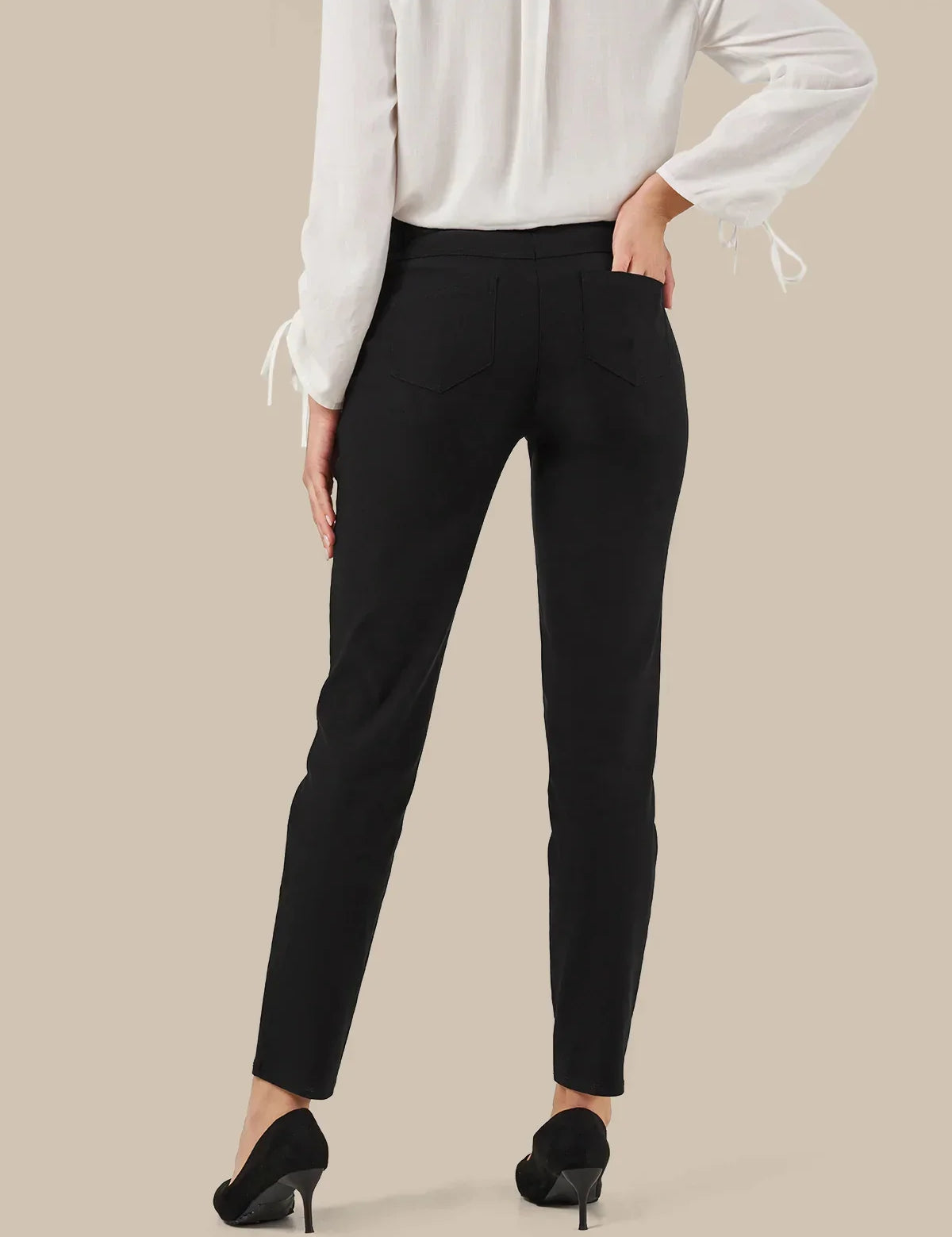A model from the back wearing slim-fit black trousers, a white long-sleeve blouse with tie details at the cuffs, and black high heels, set against a solid beige background.